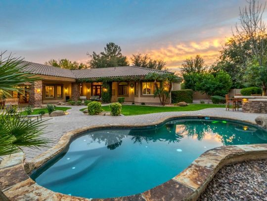 A luxurious backyard at sunset. There is a pool, an outdoor dining area, and pretty landscaping.