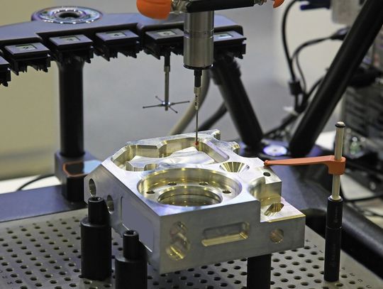 A CMM machine with multiple stylus attachment types measures a machined metal part on a fixture plate.