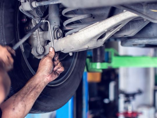 A person's hands using wrenches on a raised vehicle's rear suspension near a tire inside an auto repair shop.