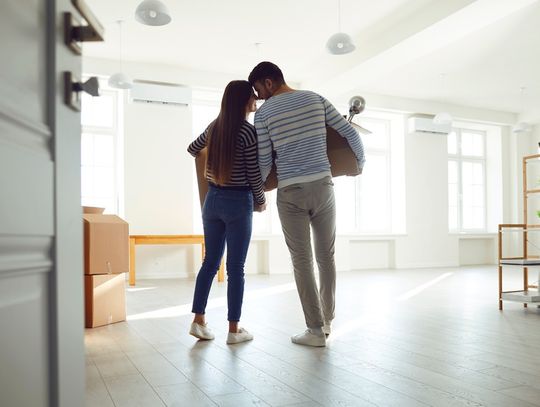 A man and a woman are standing in a room that is not furnished. They’re each holding cardboard boxes.