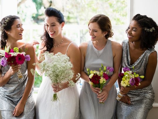 A woman in a white dress standing with three women in silver dresses holding bouquets near a bright window.