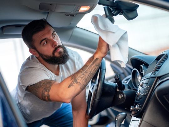 A man with tattoos is kneeling inside of a car. He’s using a fabric cloth to clean the rear-view mirror.