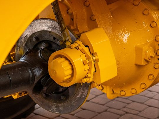 A yellow industrial machinery axle assembly with a metal brake disc, black driveshaft, and large bolted housing.