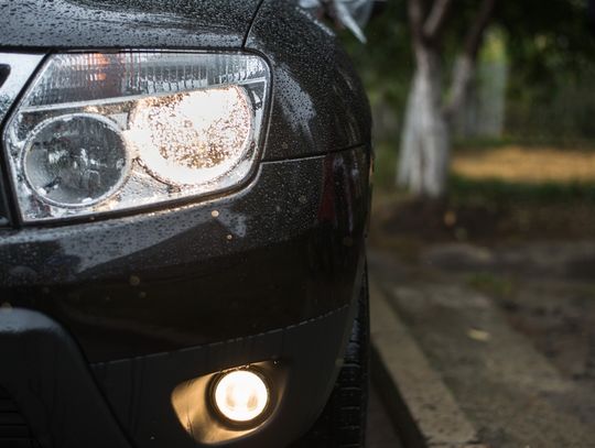 A black car with rain drops on its hood and bumper, showing an illuminated headlight and fog light outdoors.