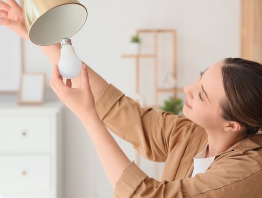 A woman in a brown shirt replacing a light bulb in a gold lamp, holding the fixture with one hand in a bright, modern home.