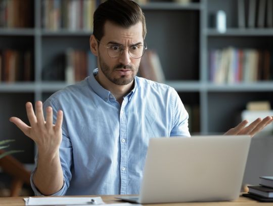 A man wearing a blue button-down shirt is raising his hands in frustration while looking at his computer.