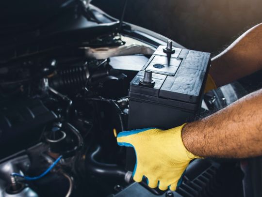 A close-up of a person holding a car battery inside an open engine compartment with tools and cables nearby.