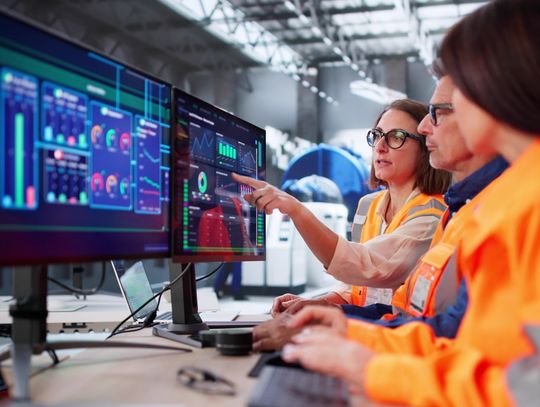 A group of three engineers meeting in front of a three screen computer setup. One of them is pointing at a screen.