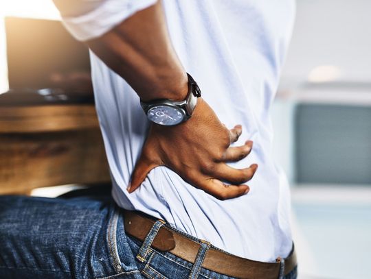 A close-up of a man wearing jeans, a brown belt, and a watch, sitting behind a desk and touching his left lower back.
