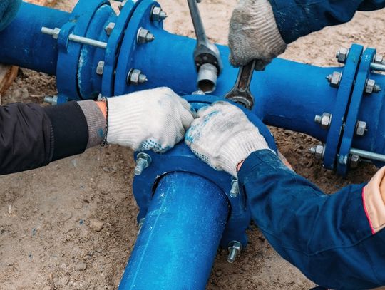 Two workers wearing gloves, loosening the bolts of a blue pipeline. One has a regular wrench and the other has a socket wrench.