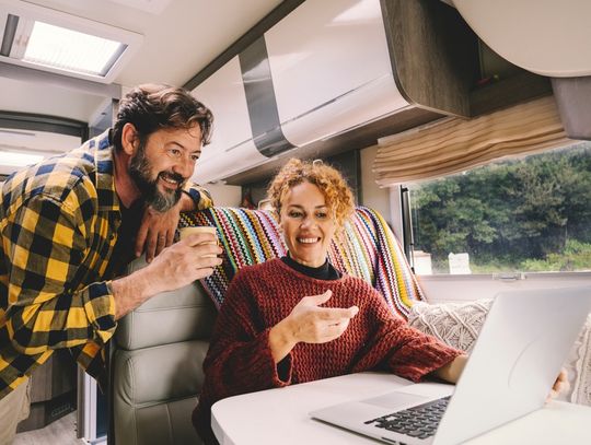 A man and a woman are sitting inside an RV. The woman is using a laptop and gesturing to the screen.