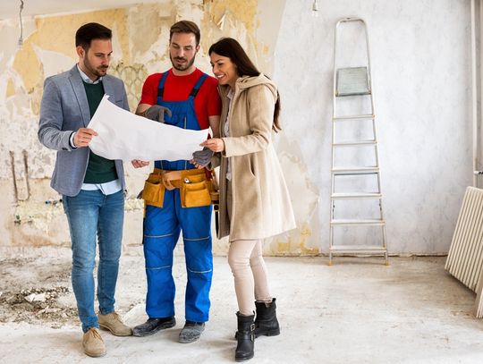 Three people looking at the blueprints for a house. They are standing in a room that is under renovation.