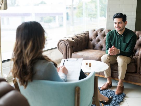 Two people are sitting across from one another. One is holding a clipboard and the other is not holding anything.