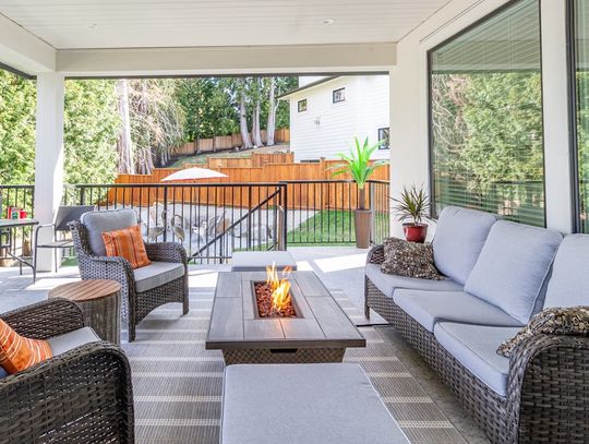 A covered patio has matching brown wicker furniture with blue-gray cushions. A fire burns in the rectangular fire pit table.