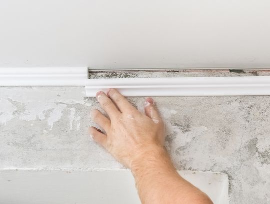 A person's hand reaches up to push crown molding into place. The molding is white and matches the ceiling.