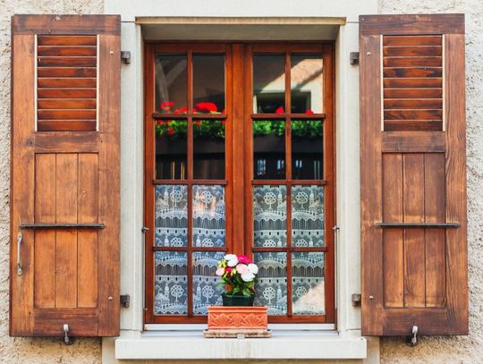 A home’s exterior window, up close. It has custom wood shutters, white lace curtains, and a small window box with flowers.