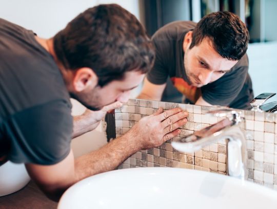 A man placing the backsplash of a sink inside a bathroom with a large mirror showing the reflection of the man.