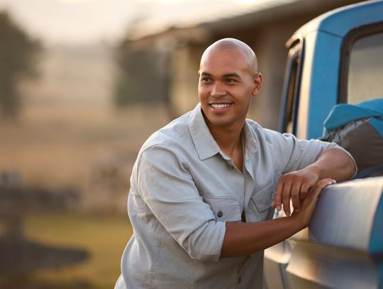 A smiling bald man leans against the left side of his blue pickup truck outside. A backpack is in the truck bed.