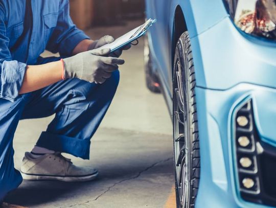 A mechanic in a blue jumpsuit kneeling next to the wheel of a blue car. He is holding a clipboard in his hand.
