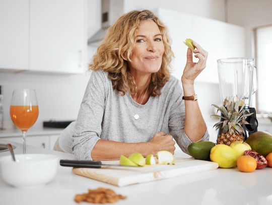A person eating a slice of fruit in their kitchen. An assortment of fruit is on the counter in front of them.