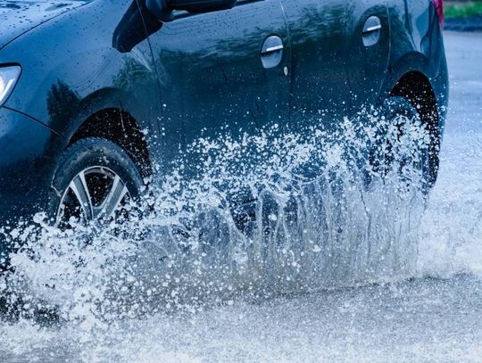 A black car is driving down the road in the rain, splashing up a big puddle of water high enough to go above the tires.