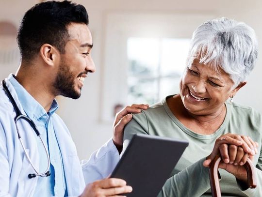 A young male doctor shows a senior female patient something on his tablet. They are smiling and happy.