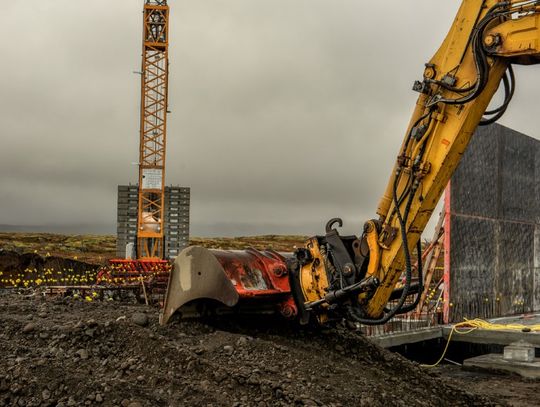 A yellow excavator lifts dark soil at a construction site, with a crane and unfinished building in the background.