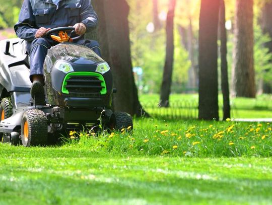 A man in a blue jumpsuit riding on a lawnmower. He is currently driving through a patch of tall grass and yellow flowers.