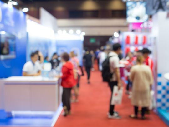 A large crowd of visitors at a trade show gather in a hallway lined with brightly lit exhibition booths.