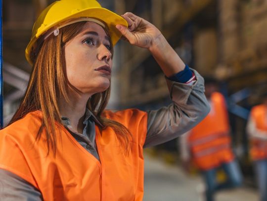 A woman wearing a yellow hard hat and an orange safety vest is standing in a warehouse. She has a hand on the brim of the hat.