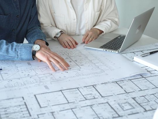 Two people are sitting at a table. There are diagrams on paper on top of the table, as well as a laptop.