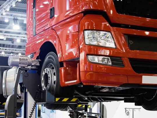 A close-up of a red truck cab resting on a car lift in a large garage, awaiting parts replacements and servicing.