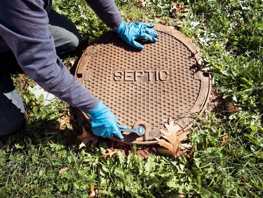 A plumber working with gloves on his hands uses a socket wrench to open a manhole cover with the word "Septic" on it.