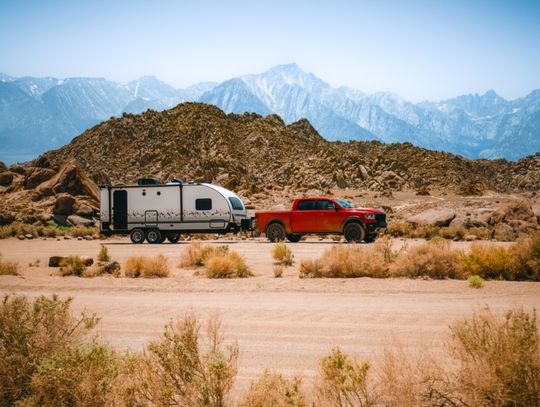 A red pickup truck hauling a travel trailer behind it. They're traveling through the desert and mountains.
