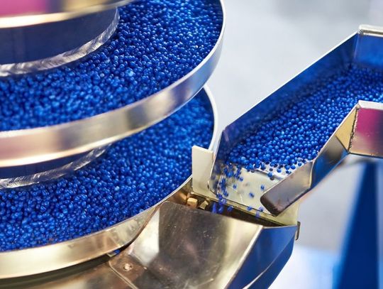 A close-up view shows thousands of tiny blue plastic pellets traveling along a stacked metal track.