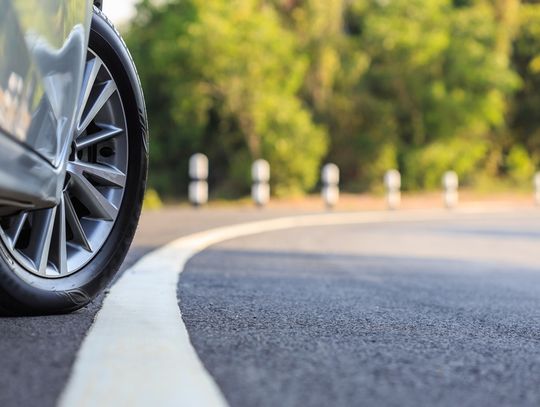 On the side of the road, a car has the front wheel turned out towards the white line. There are trees down the road.