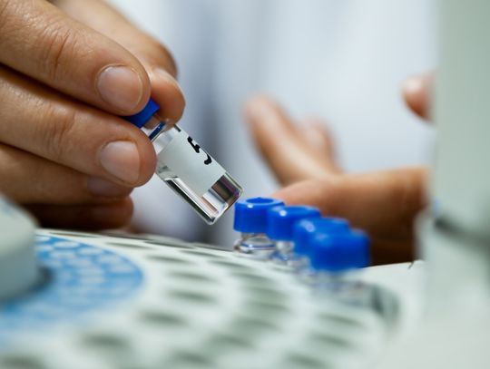 A close-up of a scientist's hand lifting a small labeled glass vial out of a chromatograph in a lab.