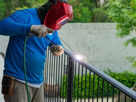 A man wearing a welding helmet and gloves working on welding the steel frame of a black fence outdoors.