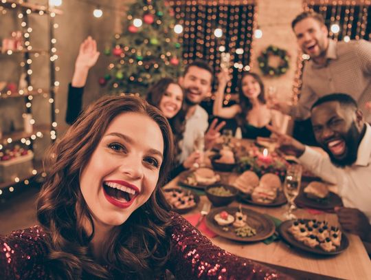 A woman takes a selfie with her party guests in the photo behind her. They're gathered for a holiday party around the table.