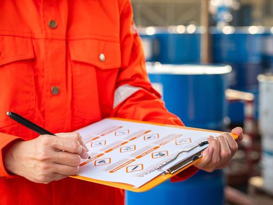 A safety officer in a bright orange jumpsuit holds and consults a checklist. Several blue barrels are in the background.