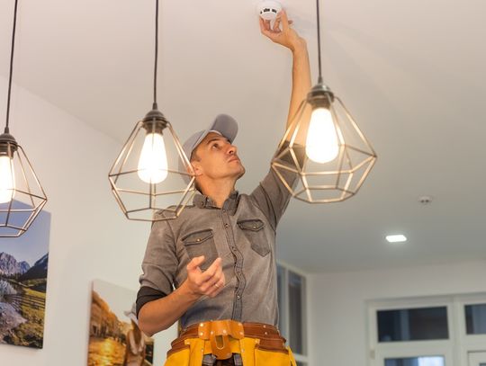 A guy wearing a gray hat and a yellow tool belt installs a new smoke detector on the ceiling of a home.