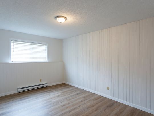 An empty room features a white baseboard heater, a wooden floor, white wood paneling, and a single window.