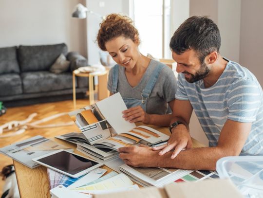 A couple sits together at a dining room table to discuss renovation details. The table contains books, plans, and swatches.
