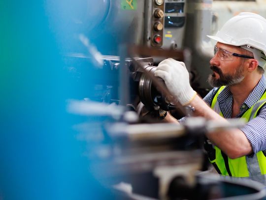 A man is wearing safety goggles, white gloves, and a hard hat as he's kneeling beside and working on a metal lathe machine.