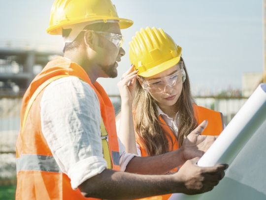 A man and a woman wearing high-visibility safety vests, hard hats, and safety goggles looking at documents outside.