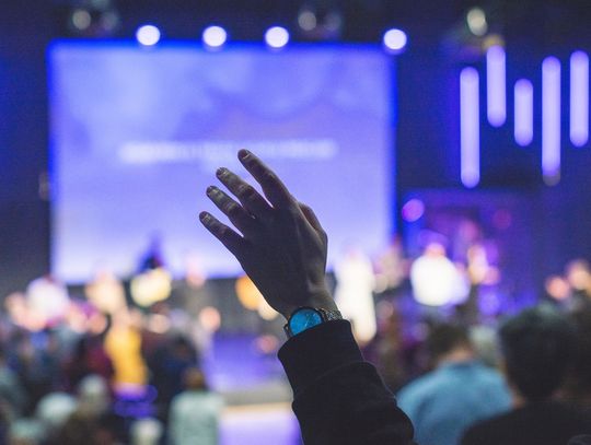 A close-up of someone's hand raised during church service. The stage lights are bright and blue as they worship.