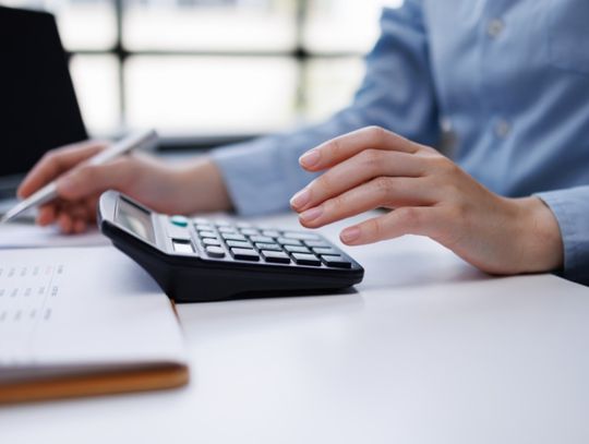Person uses a calculator while reviewing paperwork at a desk, with a pen and laptop nearby in soft focus.