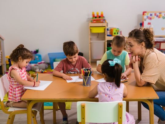 A daycare teacher guides five young children drawing at a classroom table with colored pencils and educational toys.