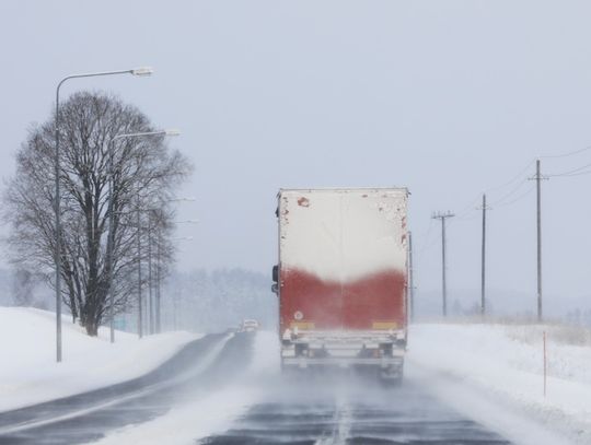 A semi truck driving on a snow-covered highway during snowfall, with trees, streetlights, and power lines along the road.