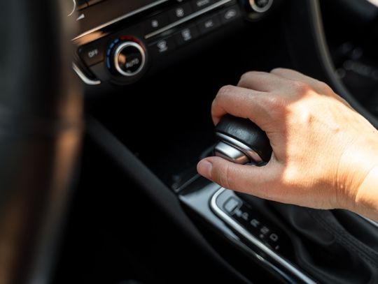 A hand holding a black gear shift in a car, with dashboard controls and sunlit center console details visible.